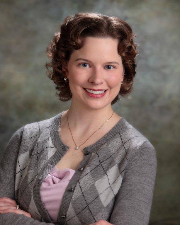 A portrait of a woman with short brown hair, wearing a gray cardigan over a pink top, smiling at the camera.